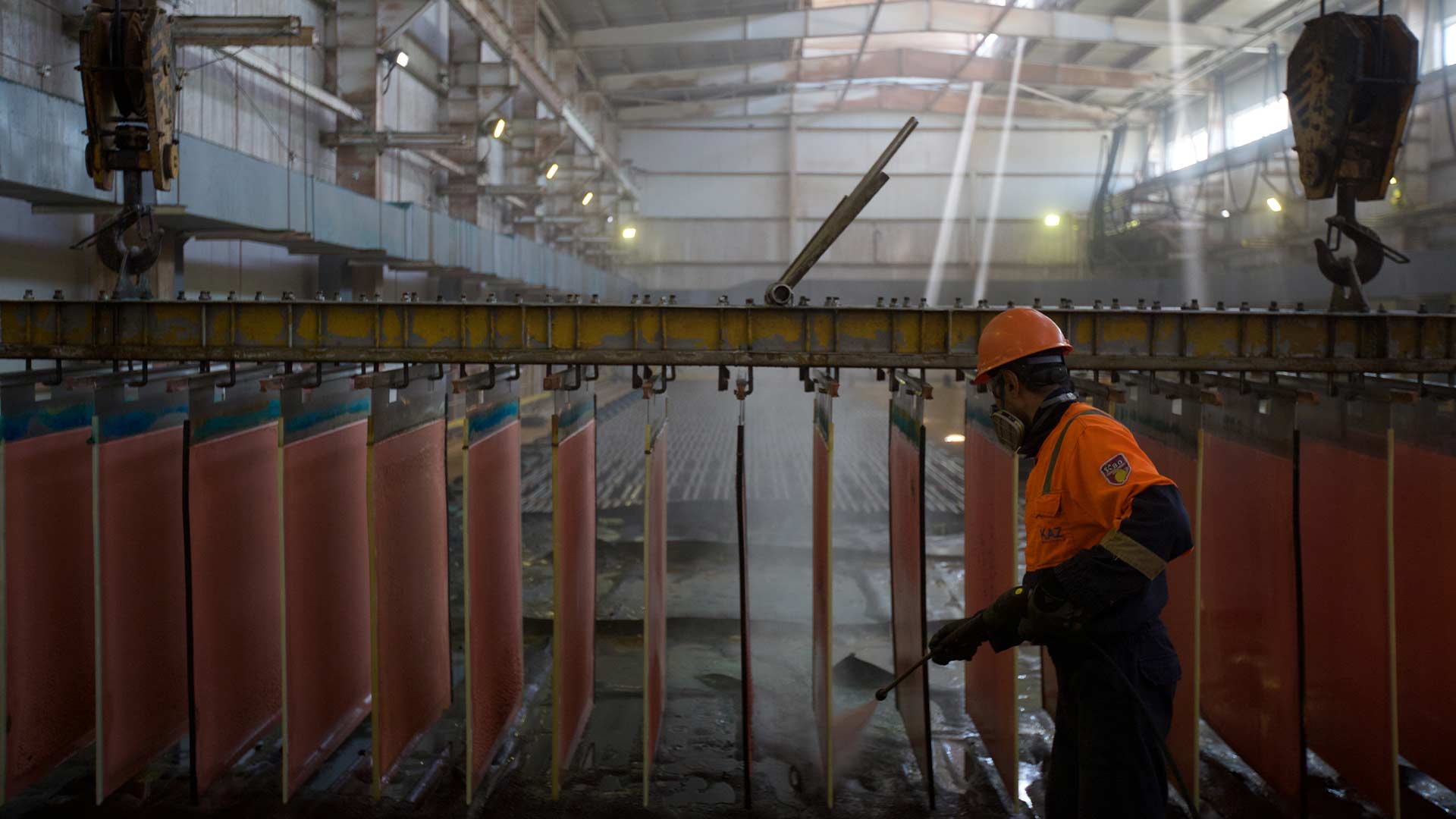 A worker in protective clothing and headgear is spraying multiple hanging sheets of copper. 