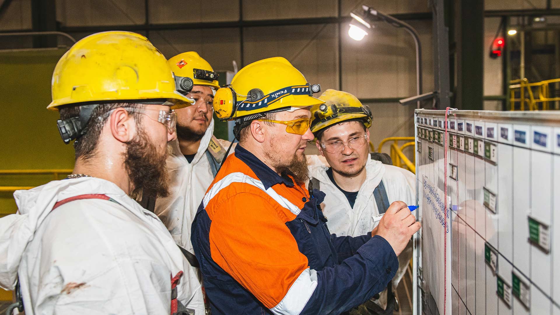 Four workers in safety clothing and protective headwear gather around a white board. One of the four is writing on the whiteboard while the other three watch. 