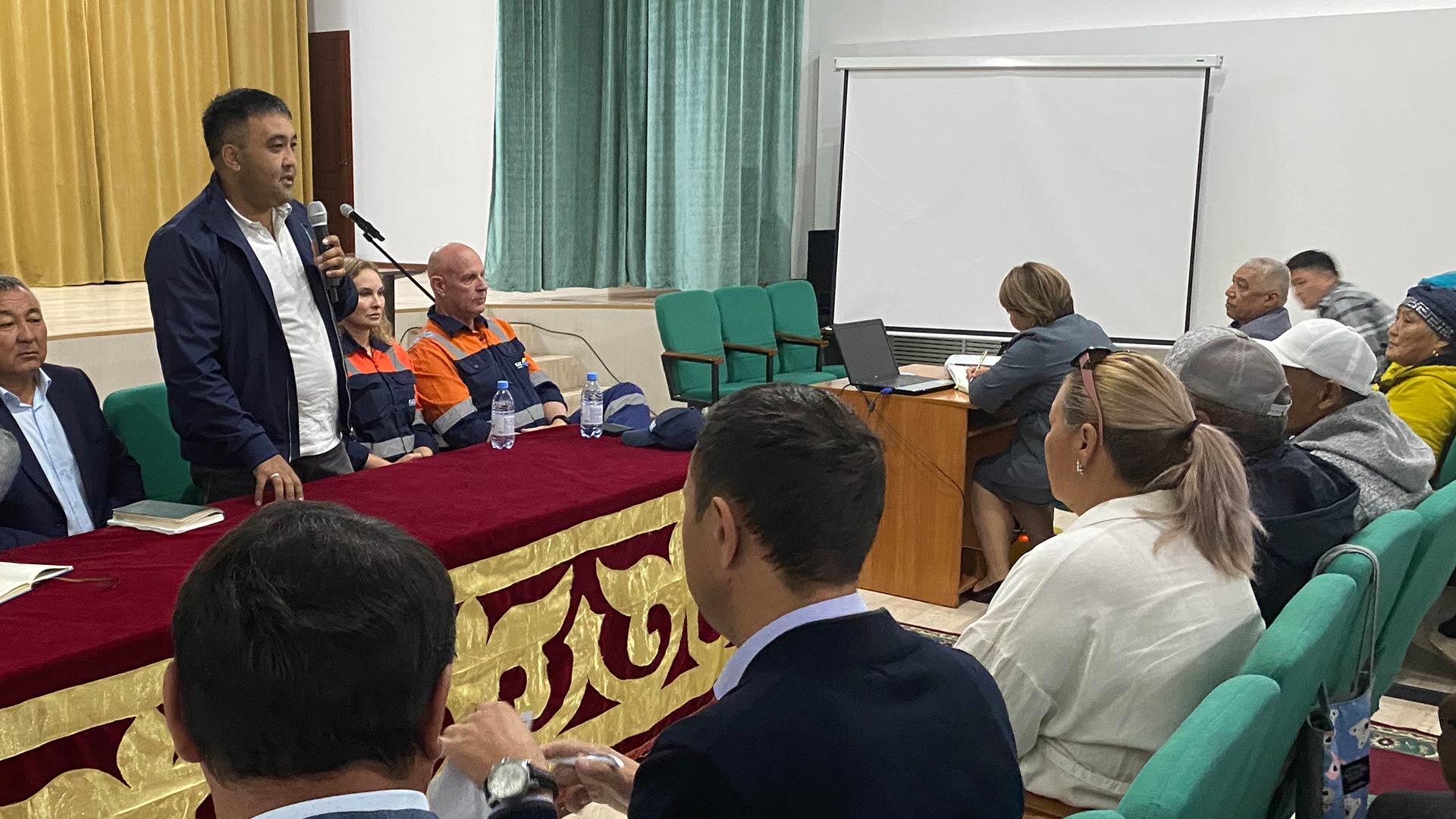 Person standing and speaking to a seated, engaged crowd.