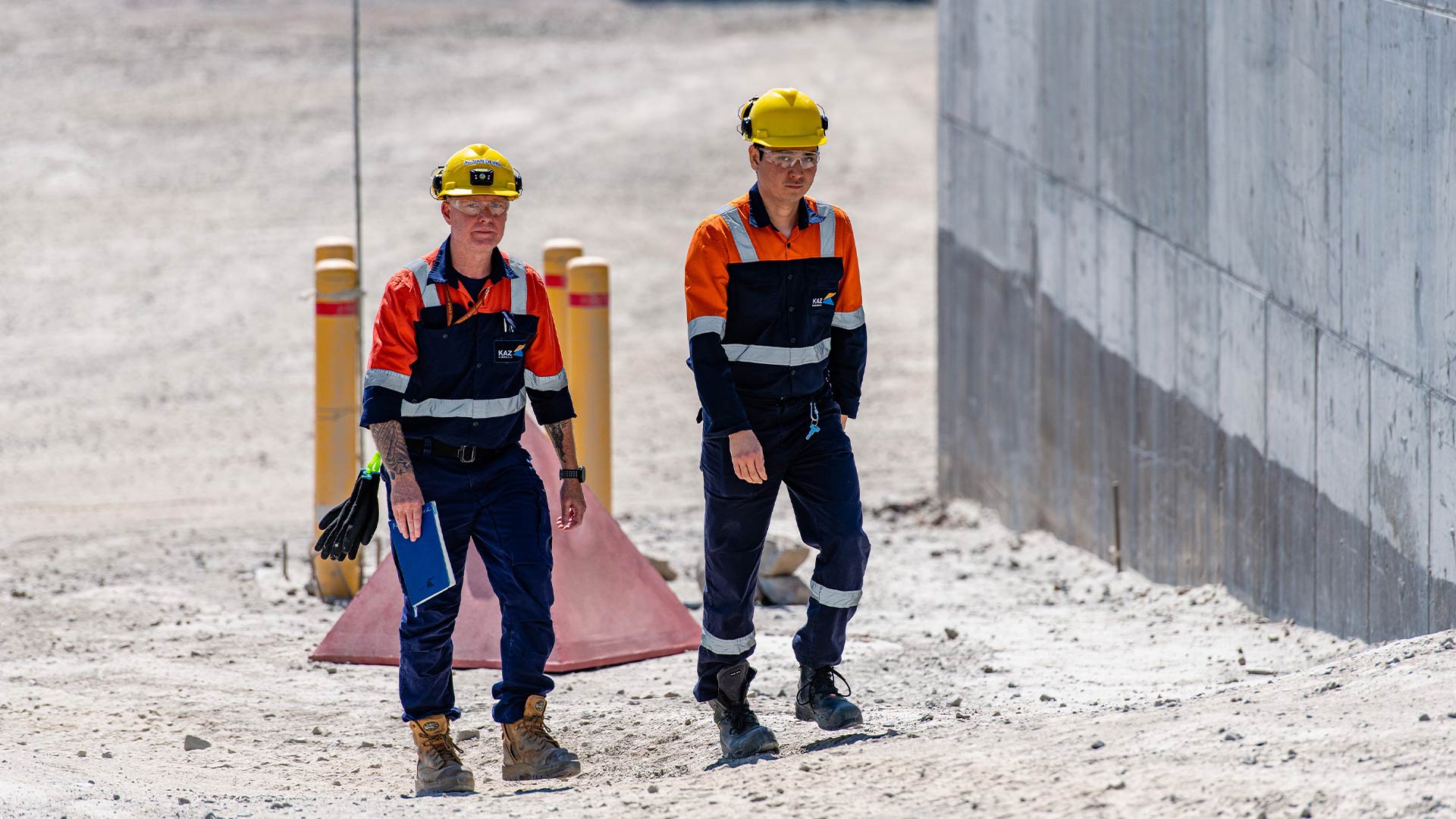 Two workers wearing protective clothing and hardhats walking up small hill. 