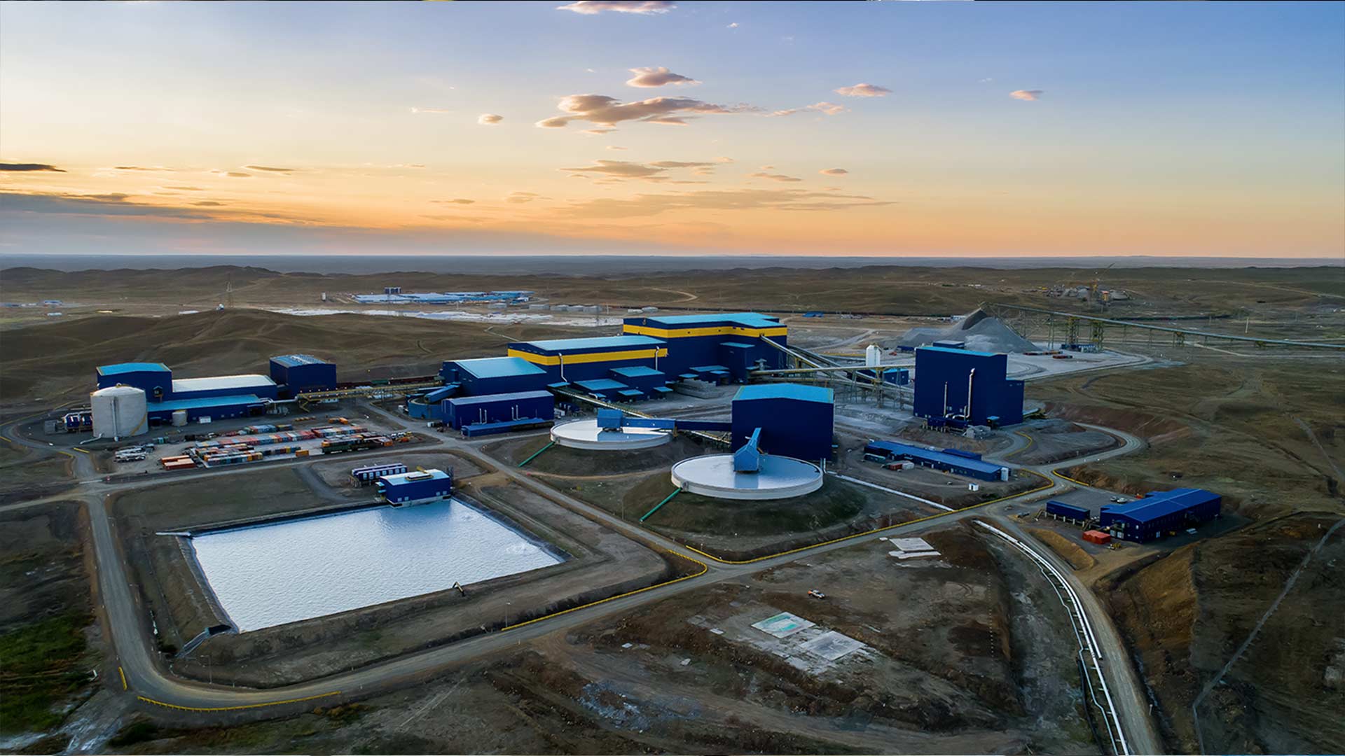 Aerial view of a vast industrial mining facility with blue buildings and large water reservoirs, set against a barren landscape at sunset.