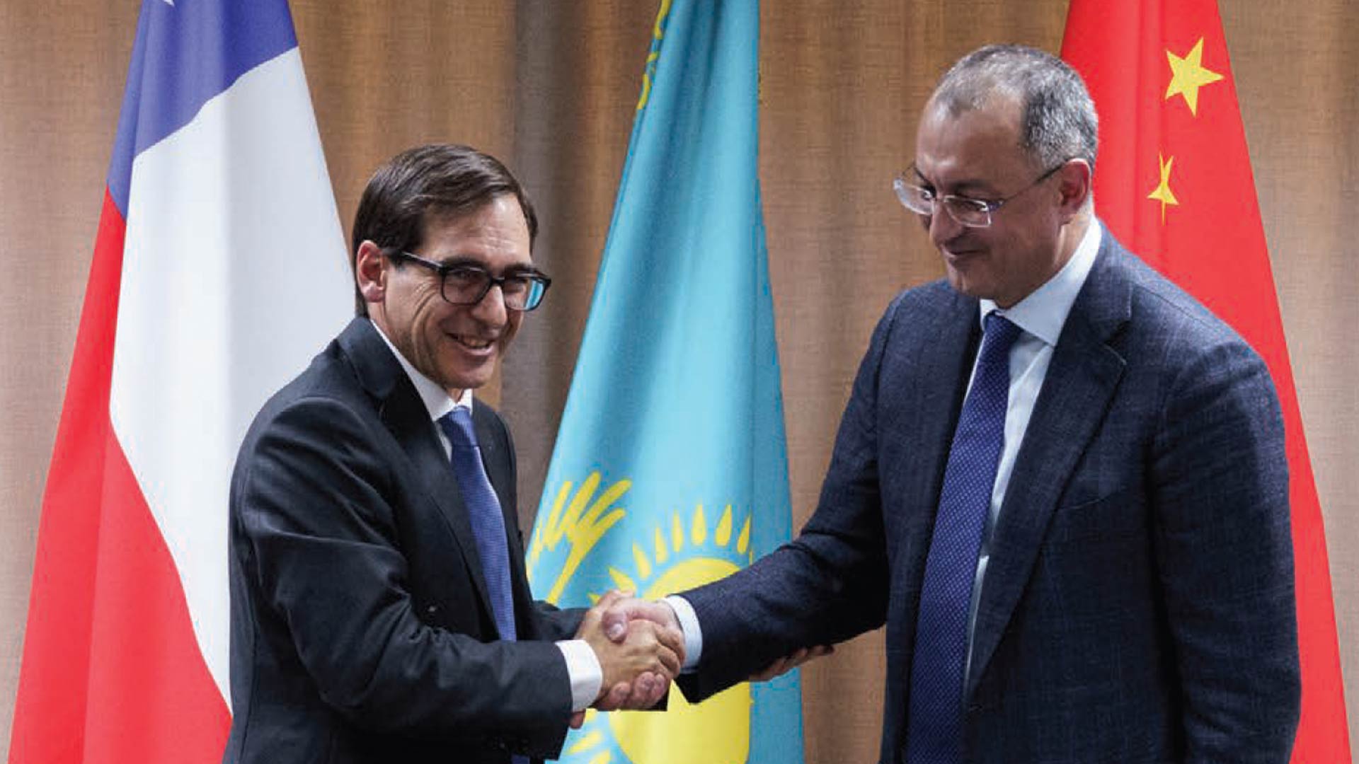 Two men in suits warmly shake hands, standing in front of flags from different countries. The atmosphere is formal and diplomatic.