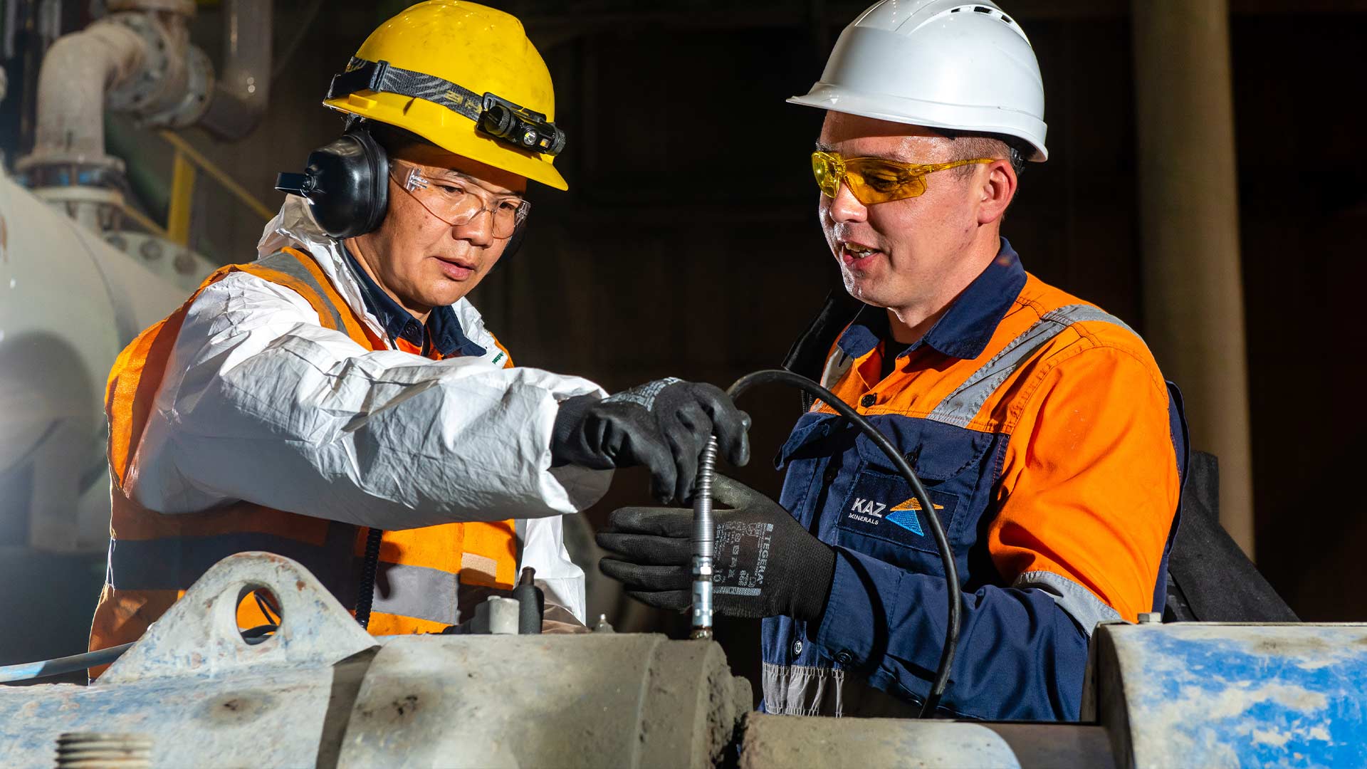 Two workers wearing safety gear inspect machinery in an industrial setting. Both wear helmets, gloves, and high-visibility vests, engaging attentively.
