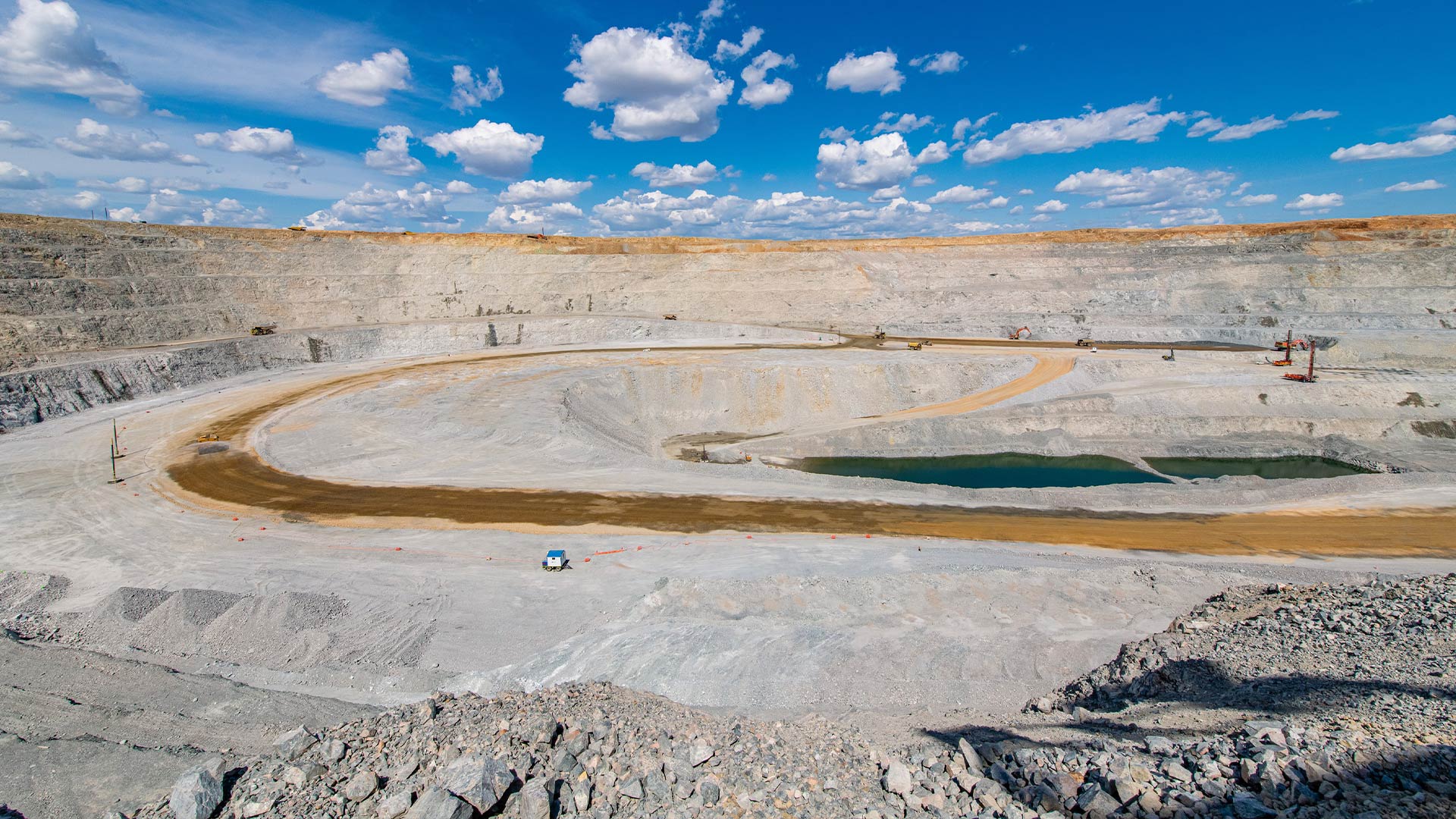 A vast open-pit mine under a bright blue sky with scattered clouds. The scene includes terraced rock walls, a winding dirt road, and a reflective pool.
