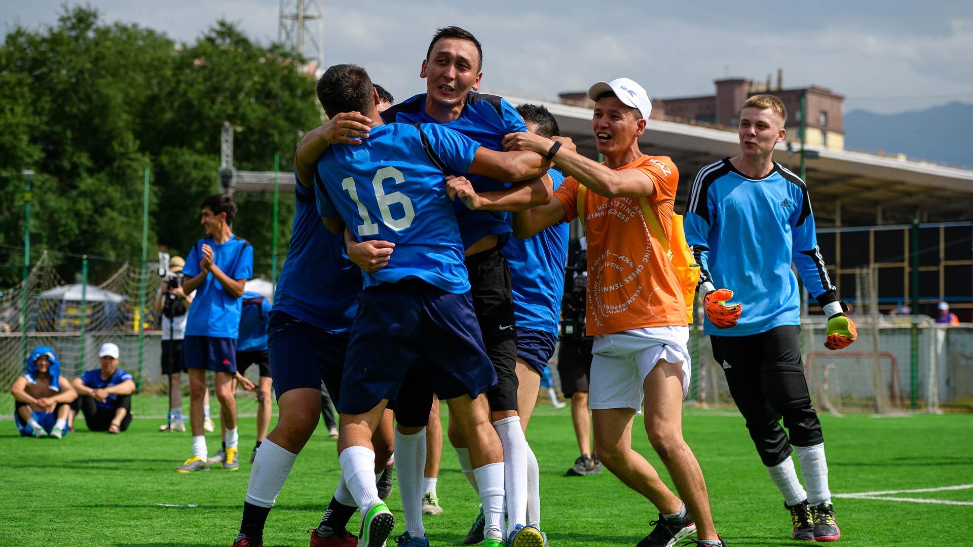Soccer team in blue and orange jerseys celebrating a win on the field. Players embrace joyfully, conveying excitement and unity under a cloudy sky.