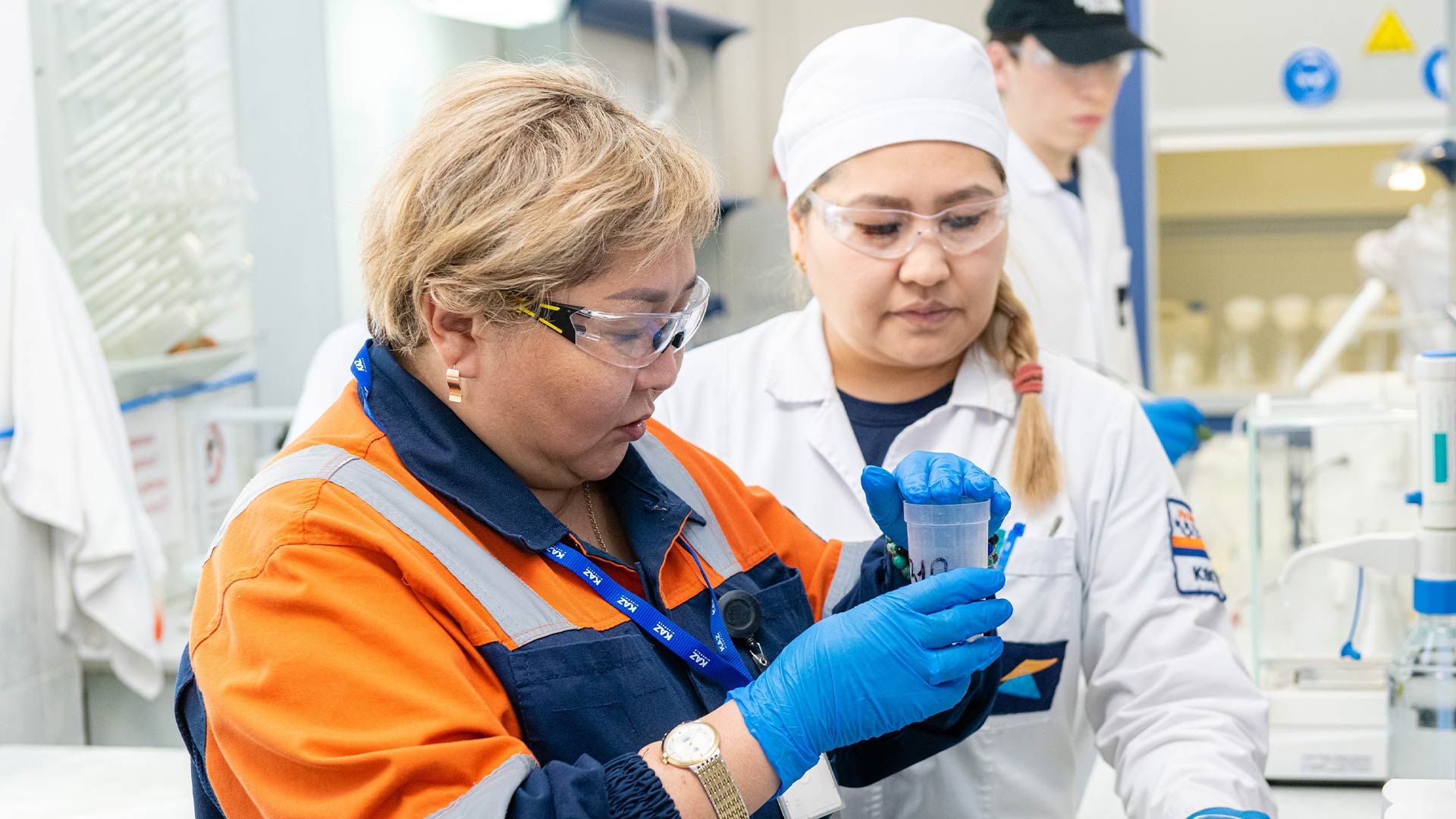 Two scientists in safety gear and gloves examine a sample in a lab. One wears an orange jacket, the other a white coat, conveying focus and teamwork.