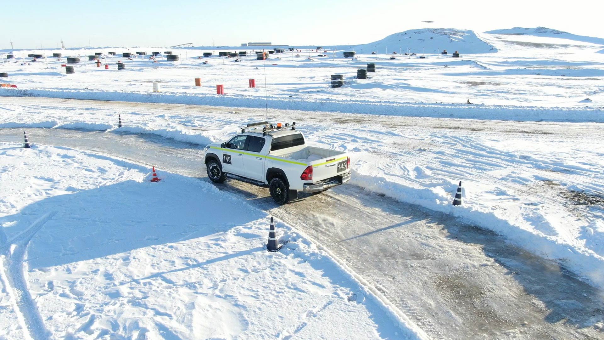 A white utility truck navigates a snowy, icy road surrounded by snow-covered landscape. Traffic cones line the path under a clear, bright sky.